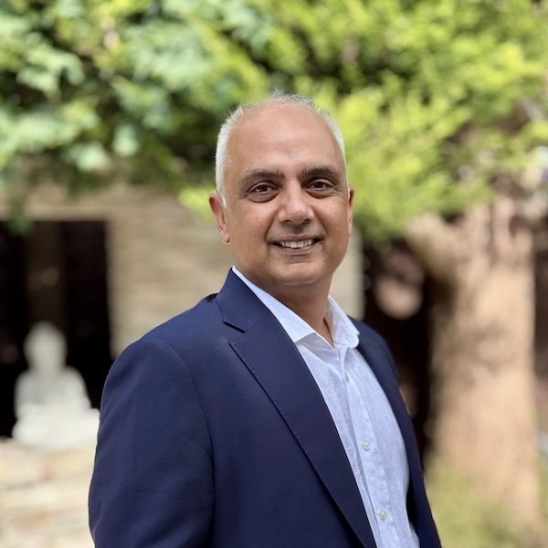 Author Arvind Rajput Smiling man in a suit standing outdoors near greenery with a serene background.